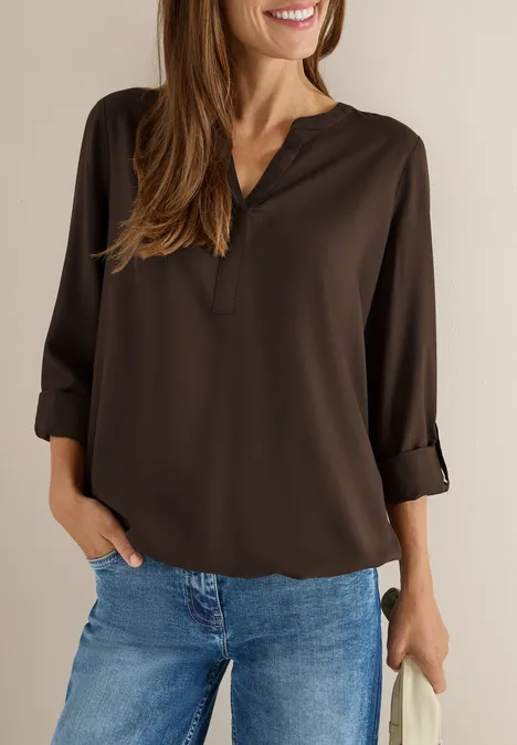 Frau mit brauner Bluse und blauer Jeans, lächelt und steht an einer Wand.